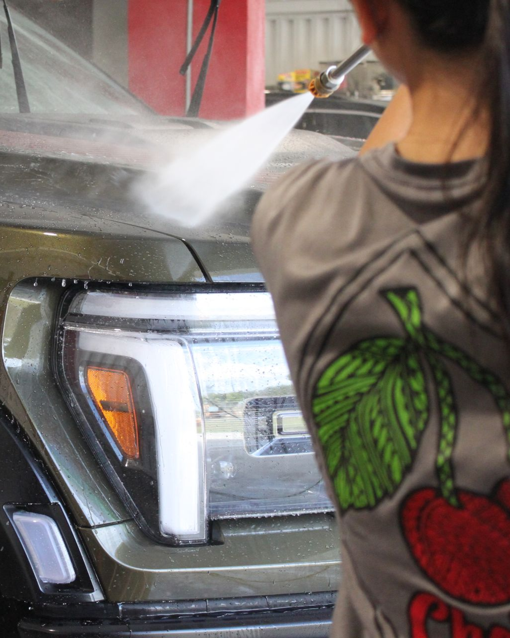 A person in a cherry-patterned shirt uses a high-pressure sprayer to wash a dark-colored vehicle at a car wash.