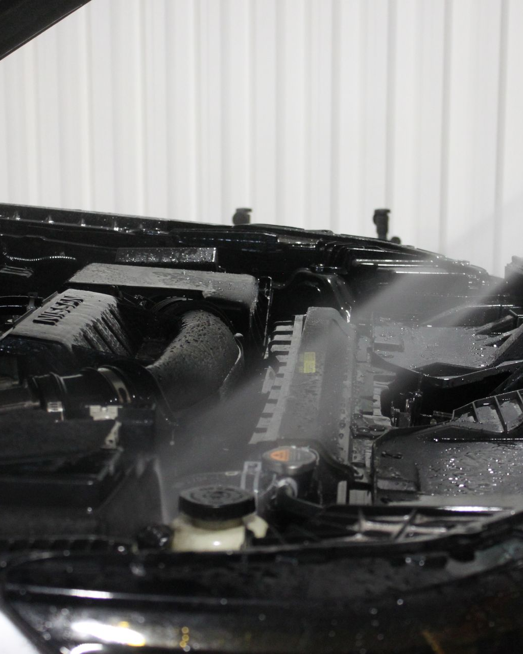 A high-pressure water spray cleans a black car engine bay in a bright, indoor facility.