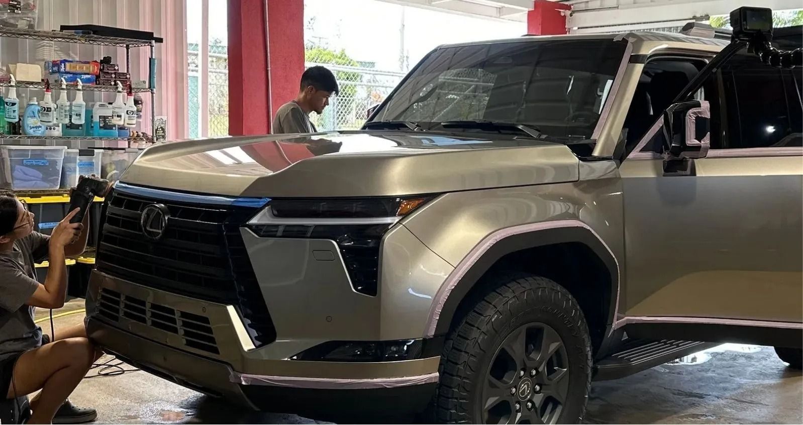 Two people in a garage working on a champagne-colored Lexus GX SUV with black wheels and a rugged off-road front bumper.