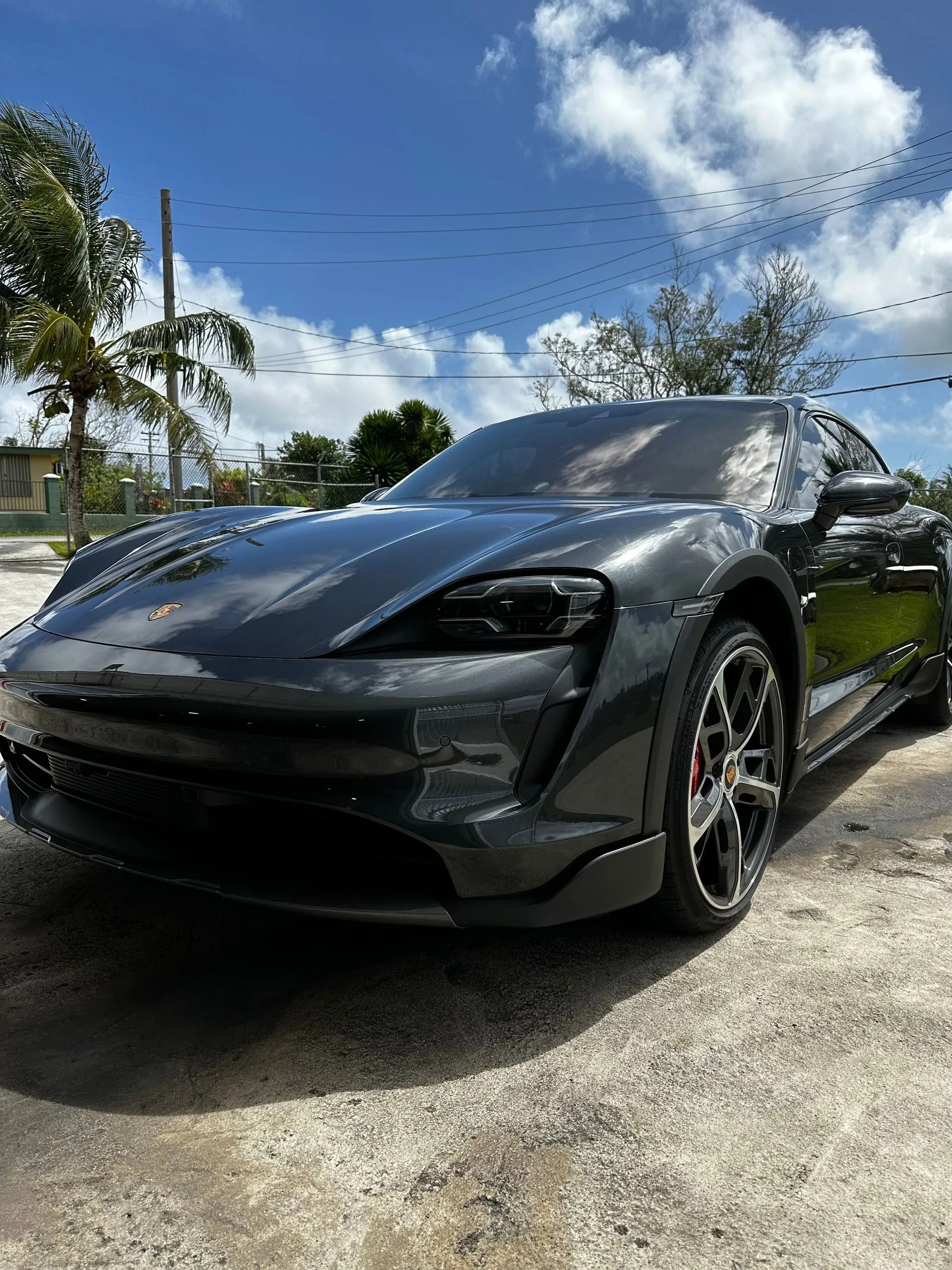 A dark gray Porsche Taycan parked on a gravel lot under a bright blue sky with scattered clouds.
