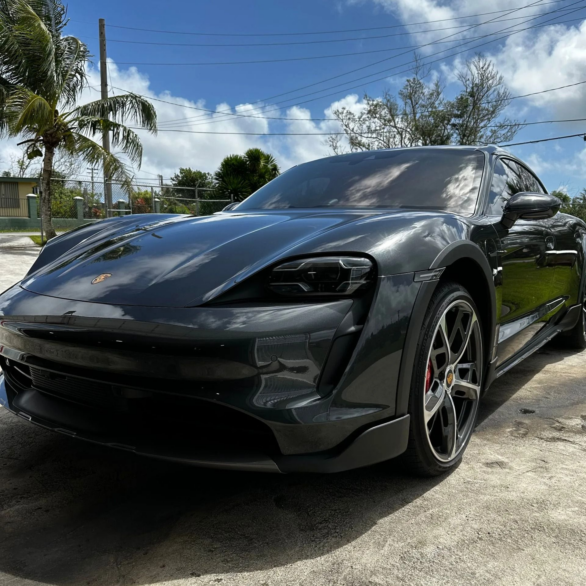 A dark grey Porsche Taycan parked on a gravel surface under a bright blue, sunny sky with palm trees in the background.