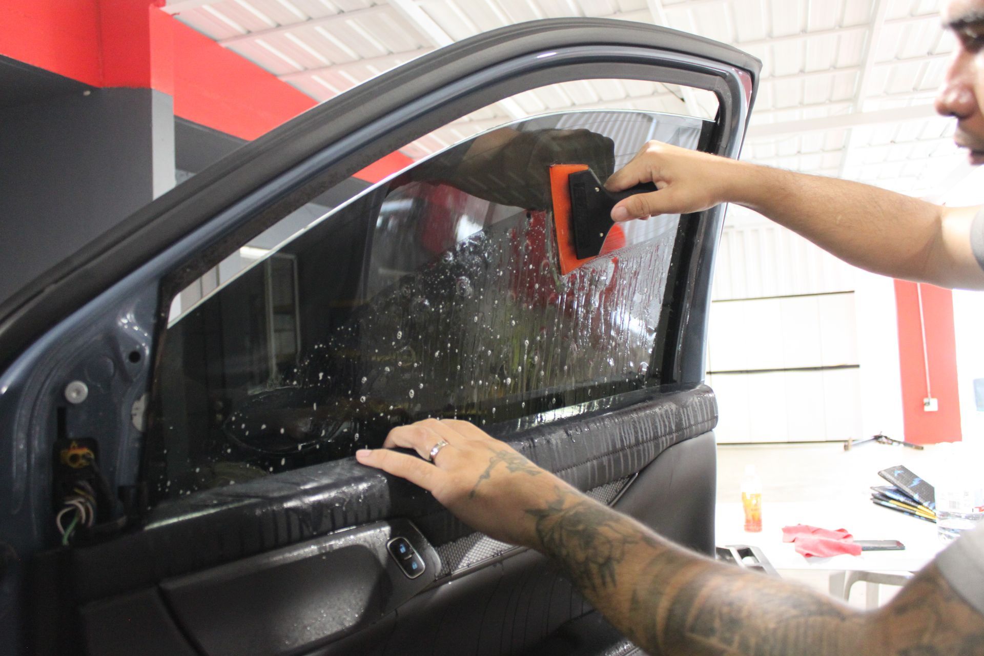 A technician uses a squeegee to apply window tint film to a car door window in a workshop.