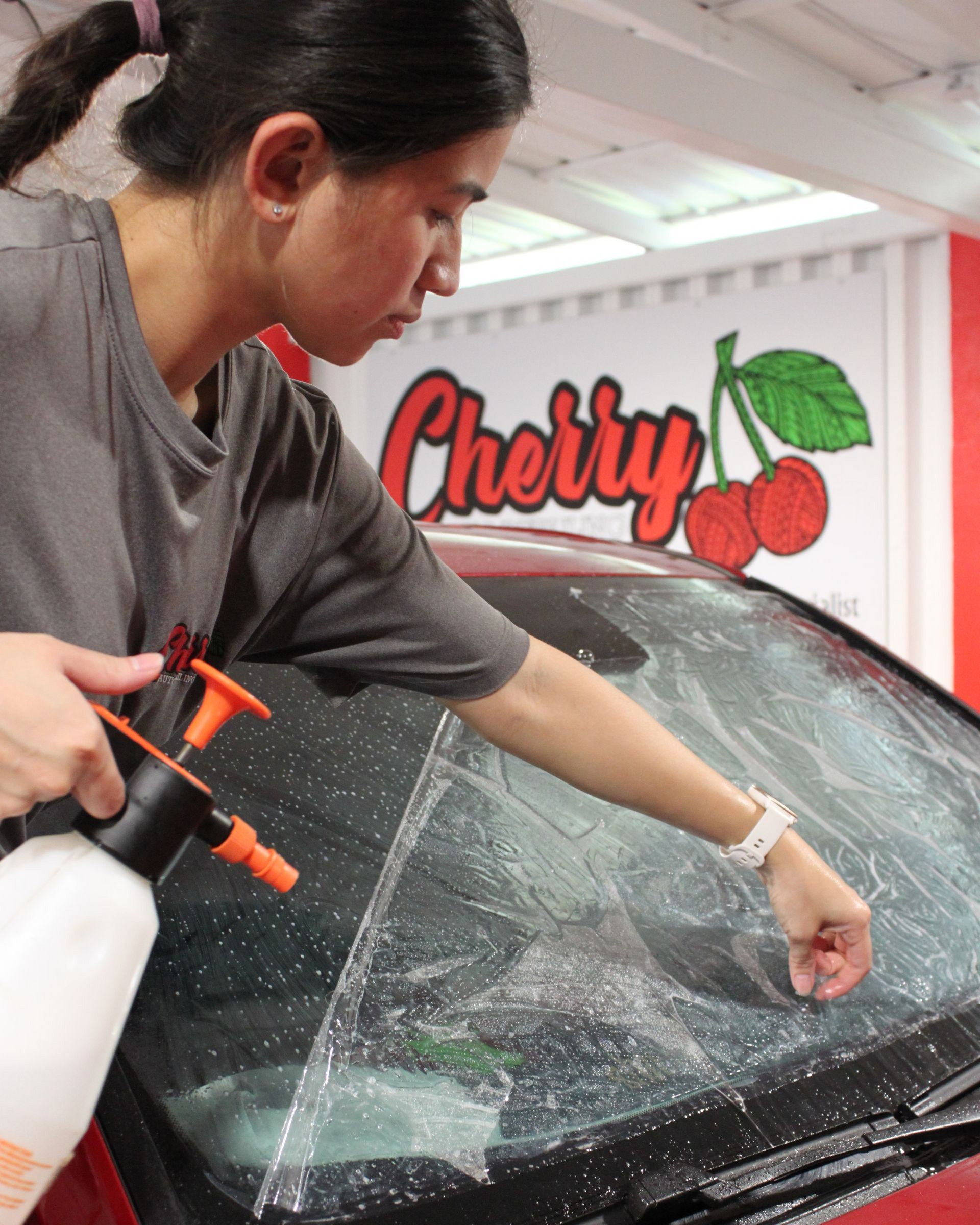 A person sprays cleaning solution onto a car windshield to apply a tint film in a shop with a cherry logo on the wall.