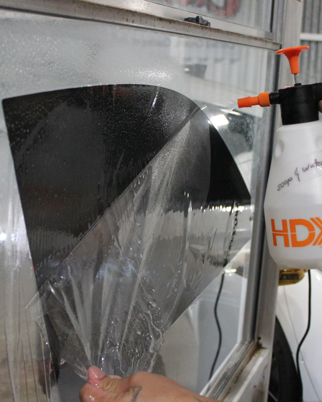 A hand peels the protective clear backing off a piece of dark window tint film, next to a spray bottle filled with liquid.