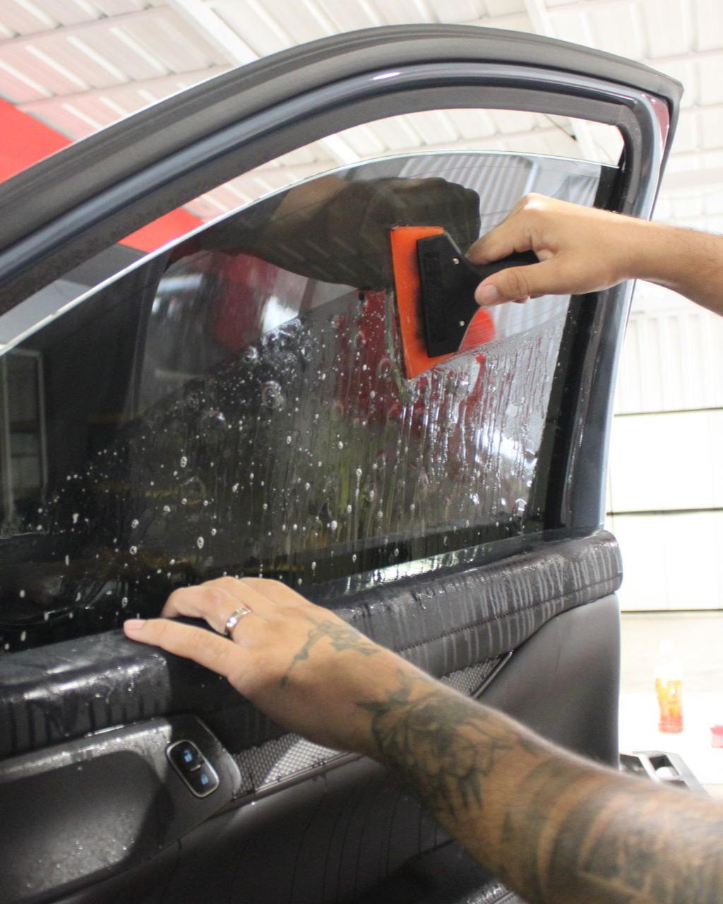 A person with tattooed arms uses an orange squeegee to smooth window tint onto a car door glass.
