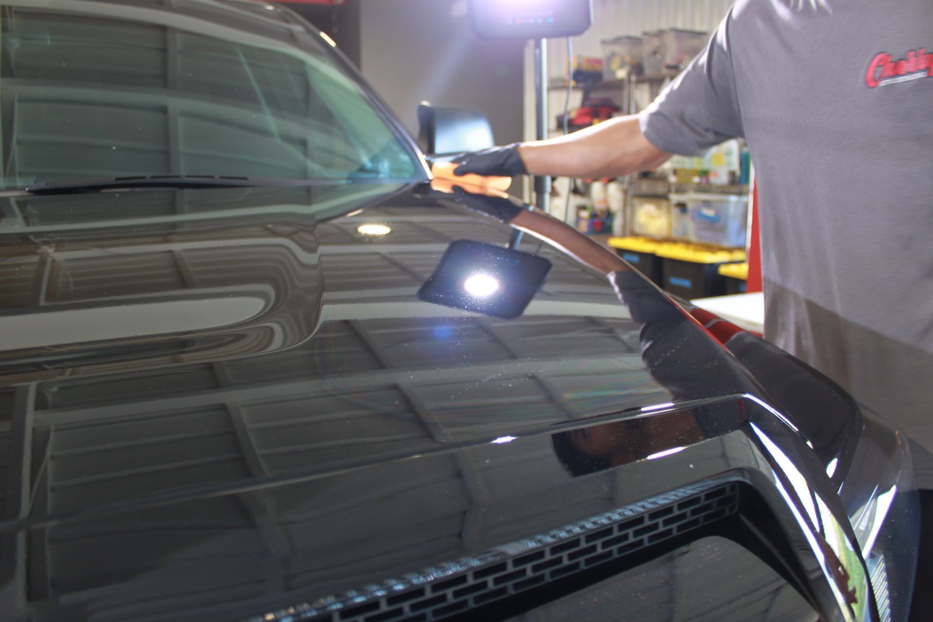 A person wearing a gray t-shirt and black glove cleans the hood of a shiny, dark-colored car in a garage.