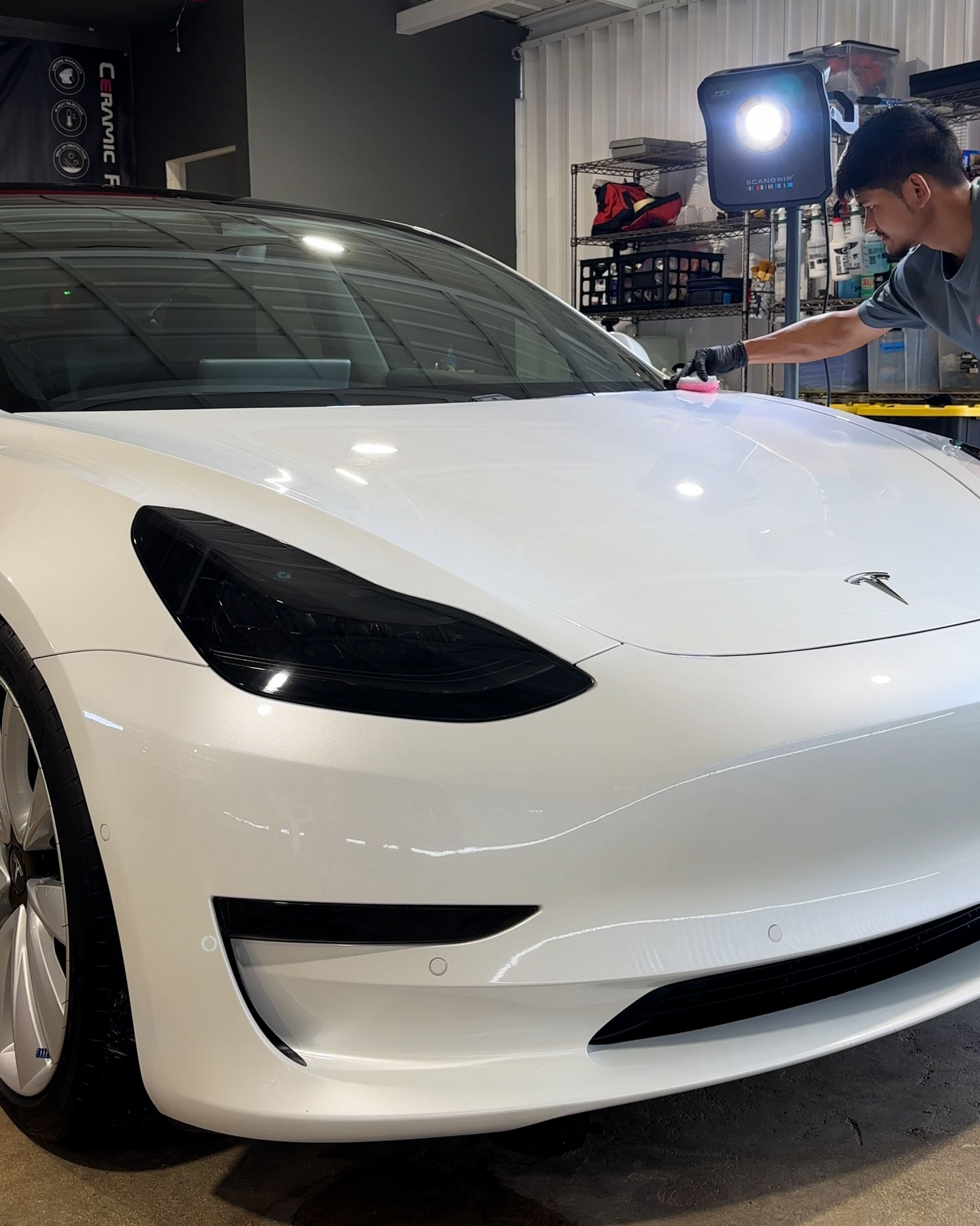 A cherry-red Tesla Model 3 sits in front of an open garage, with a matching red automotive part resting on the grass.
