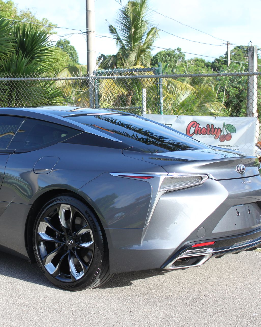 A dark gray Lexus LC coupe parked outdoors near a chain-link fence with a cherry-themed banner in the background.
