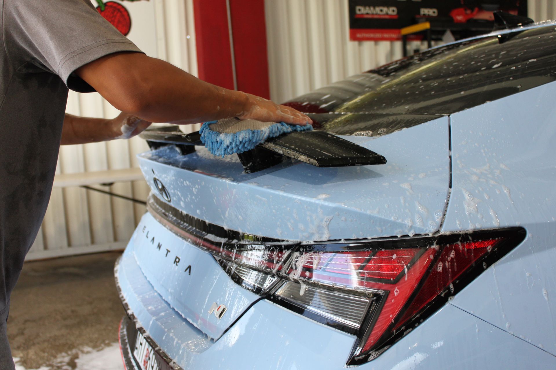 A person washes the rear spoiler and trunk of a light blue Elantra car with a soapy sponge at a car wash.