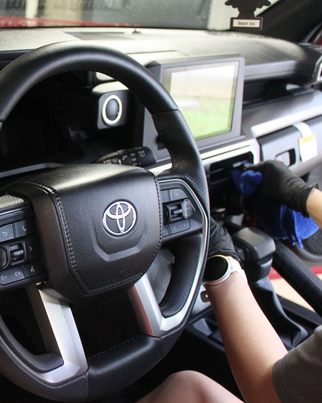A person wearing black gloves cleans the interior dashboard of a Toyota vehicle with a blue microfiber cloth.