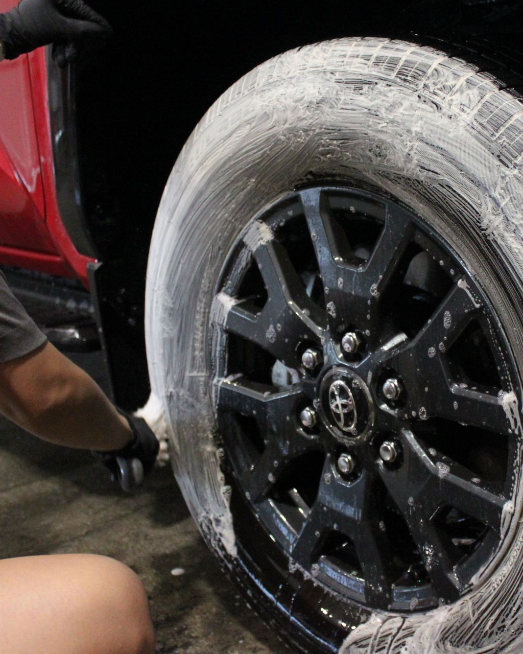 A person wearing black gloves cleans a soap-covered car tire on a black Toyota alloy wheel.