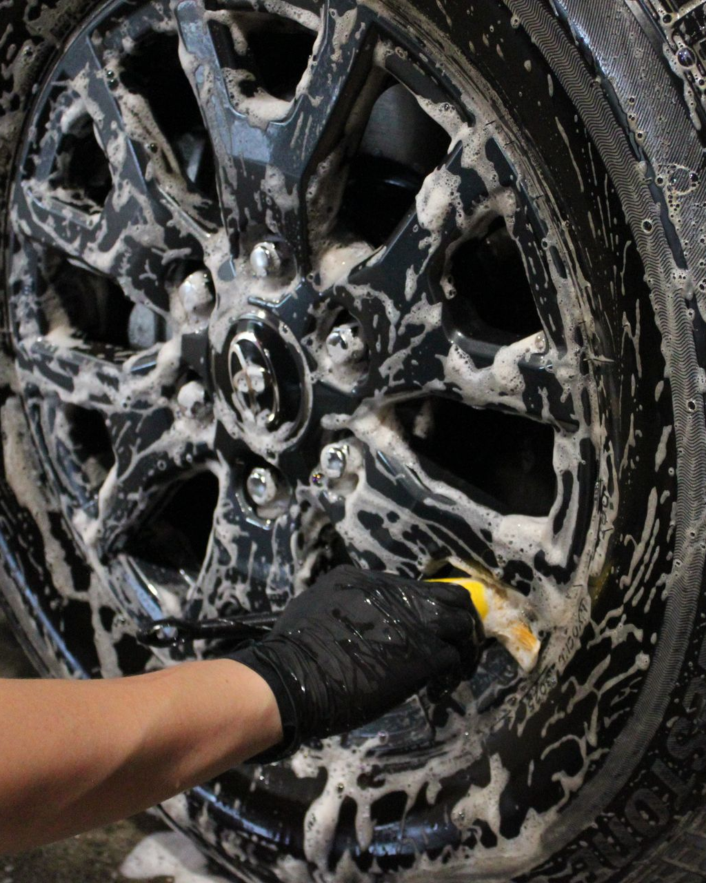 A gloved hand uses a brush to scrub foam cleaner onto a black car wheel.