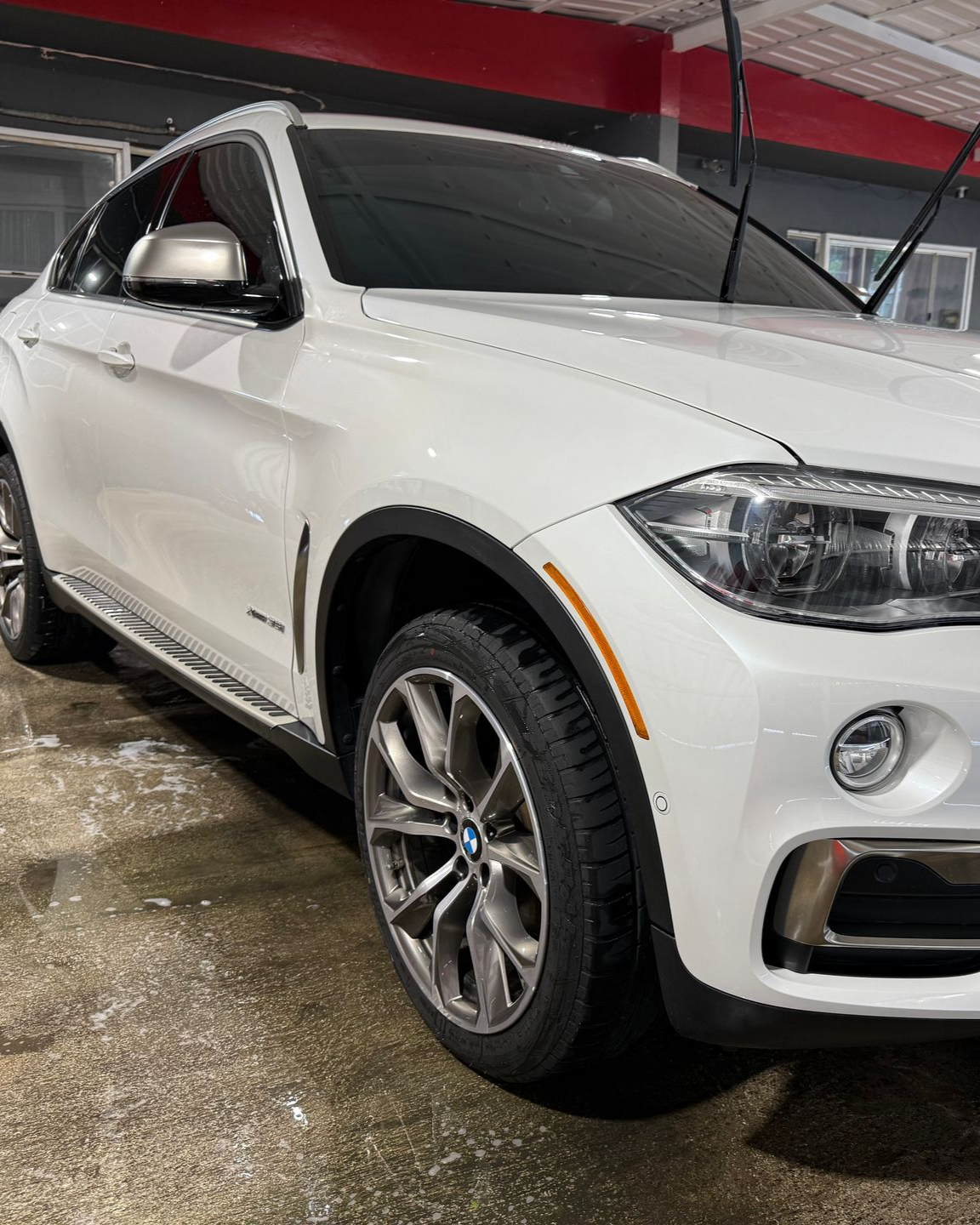 A white BMW X6 parked indoors on a wet floor, angled to show its front, passenger side, and multi-spoke wheels.
