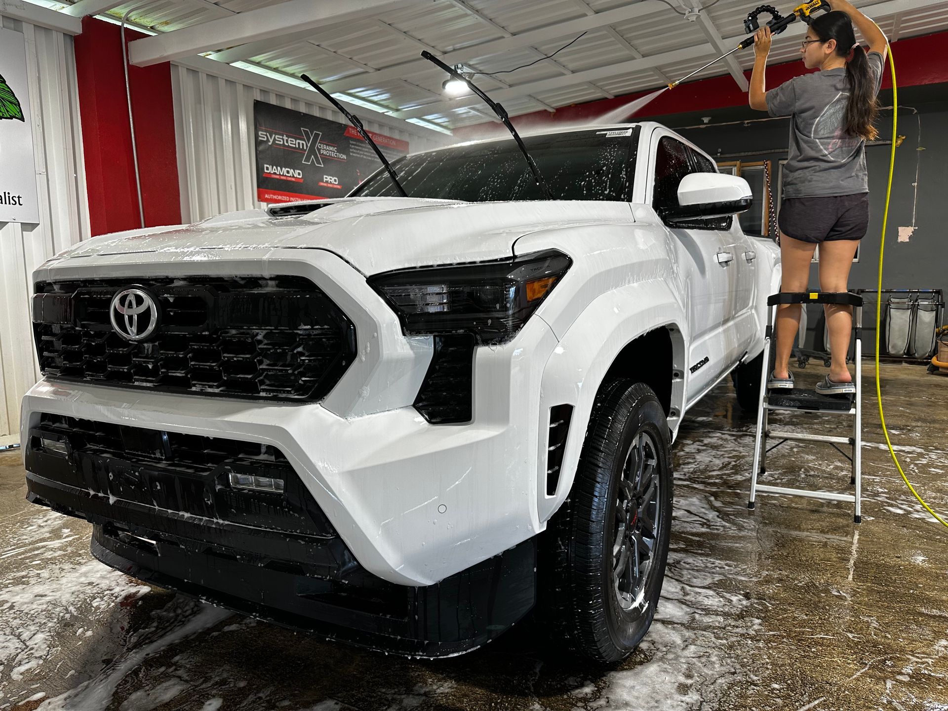 A person standing on a step ladder washes a white Toyota Tacoma truck inside a well-lit bay.