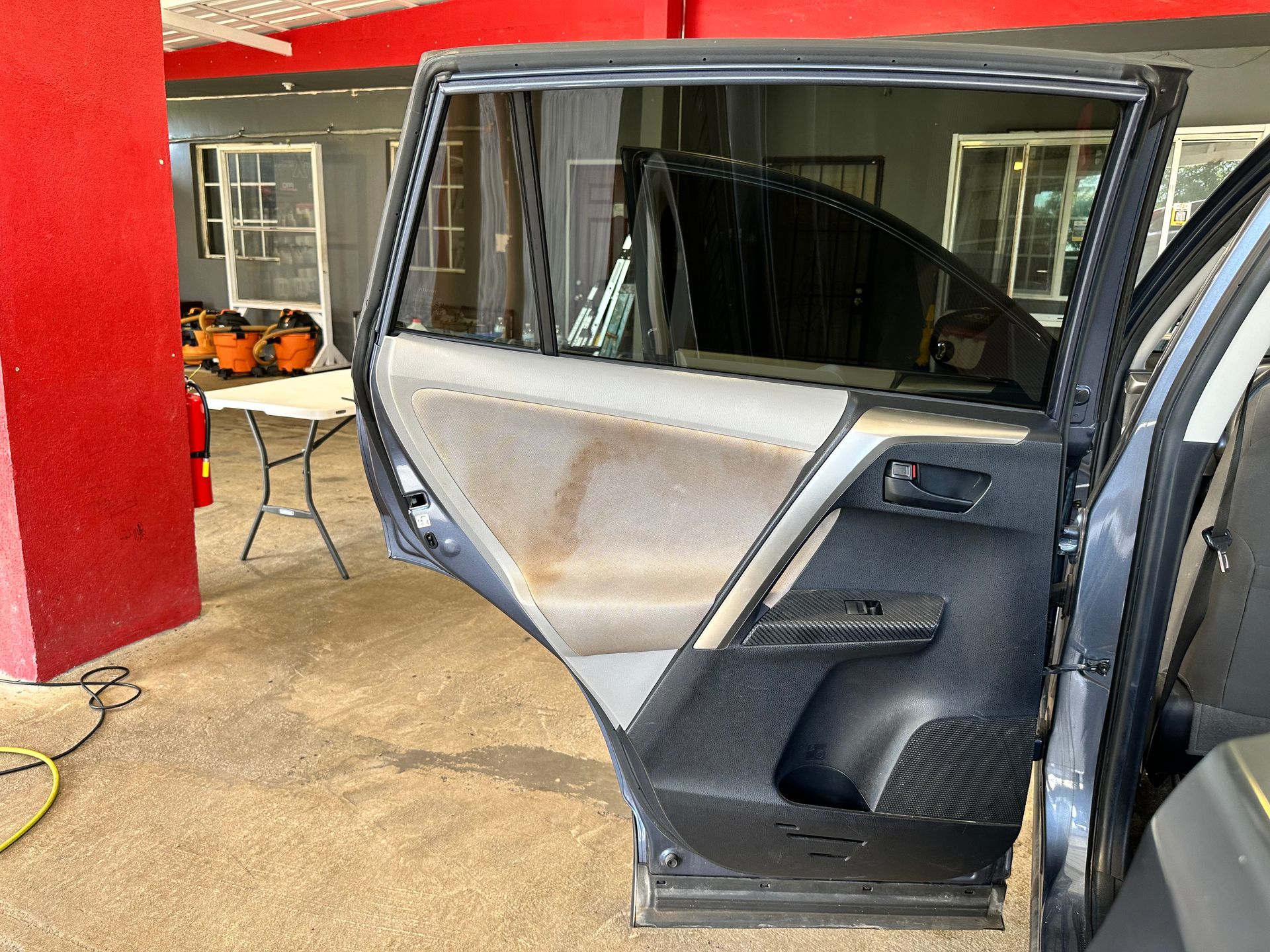 Open rear car door showing a grey fabric interior panel and a tinted window in a garage setting.