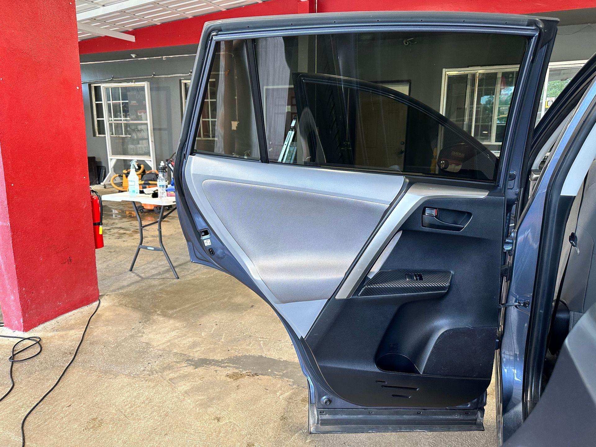 An open rear car door with a tinted window, gray fabric panel, and black trim, set in a garage with a red pillar.