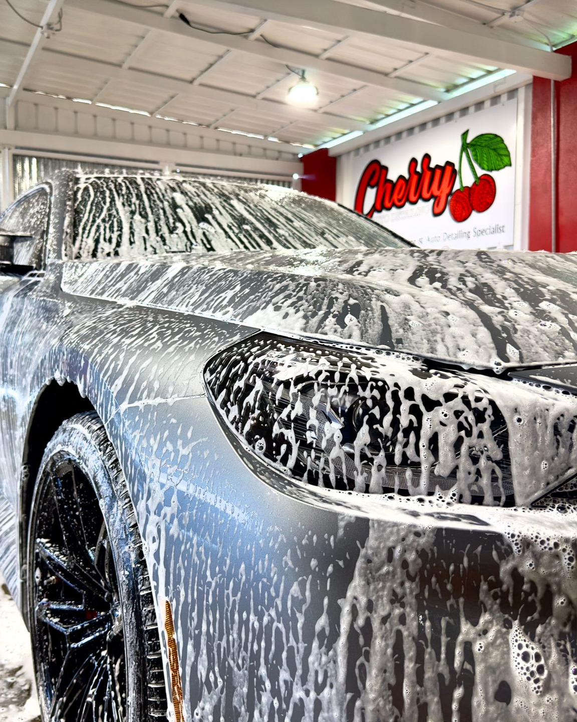 A car covered in thick white soap suds at a car wash under a sign with a cherry logo.