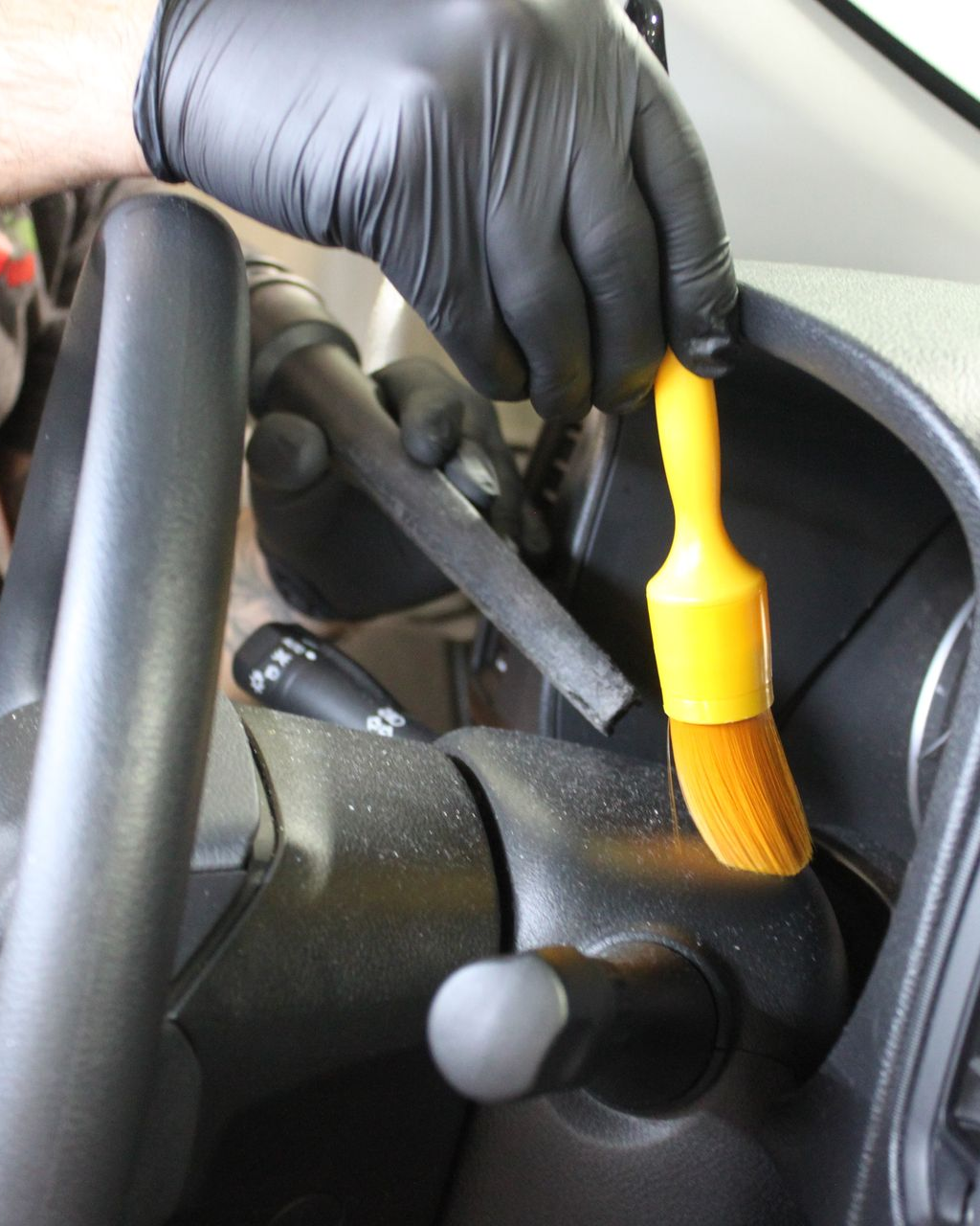 A hand in a black glove uses a yellow brush to clean dust off a car's steering column and dashboard.