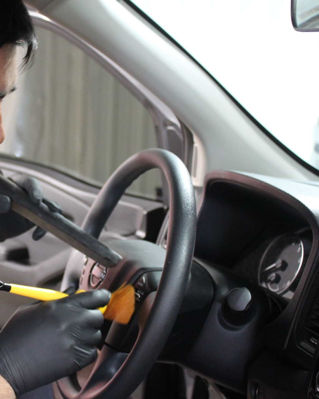 A person wearing black gloves uses a small yellow-bristled brush to clean the buttons on a car steering wheel.