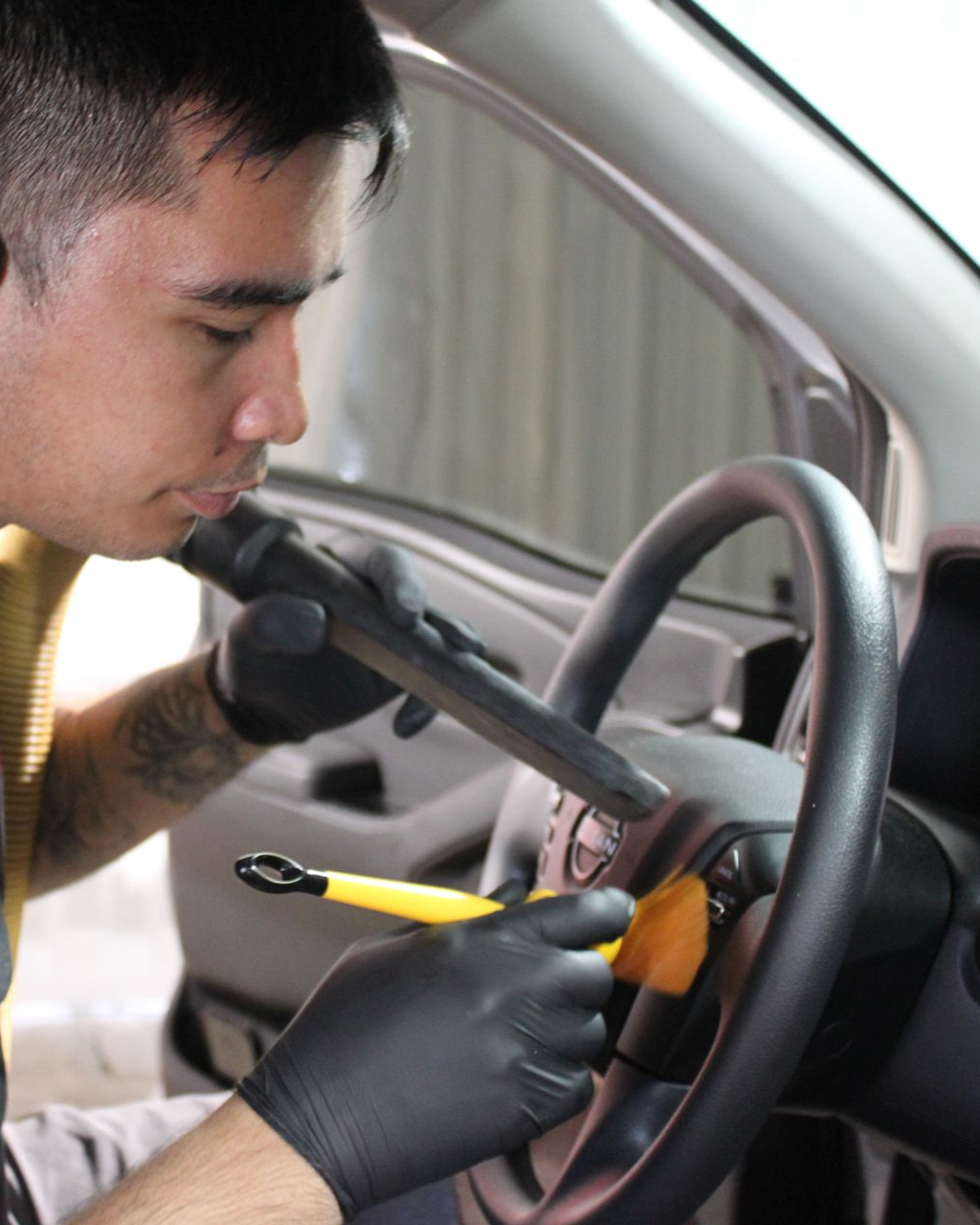 A person wearing black gloves uses a vacuum nozzle and a soft brush to clean the steering wheel of a car.