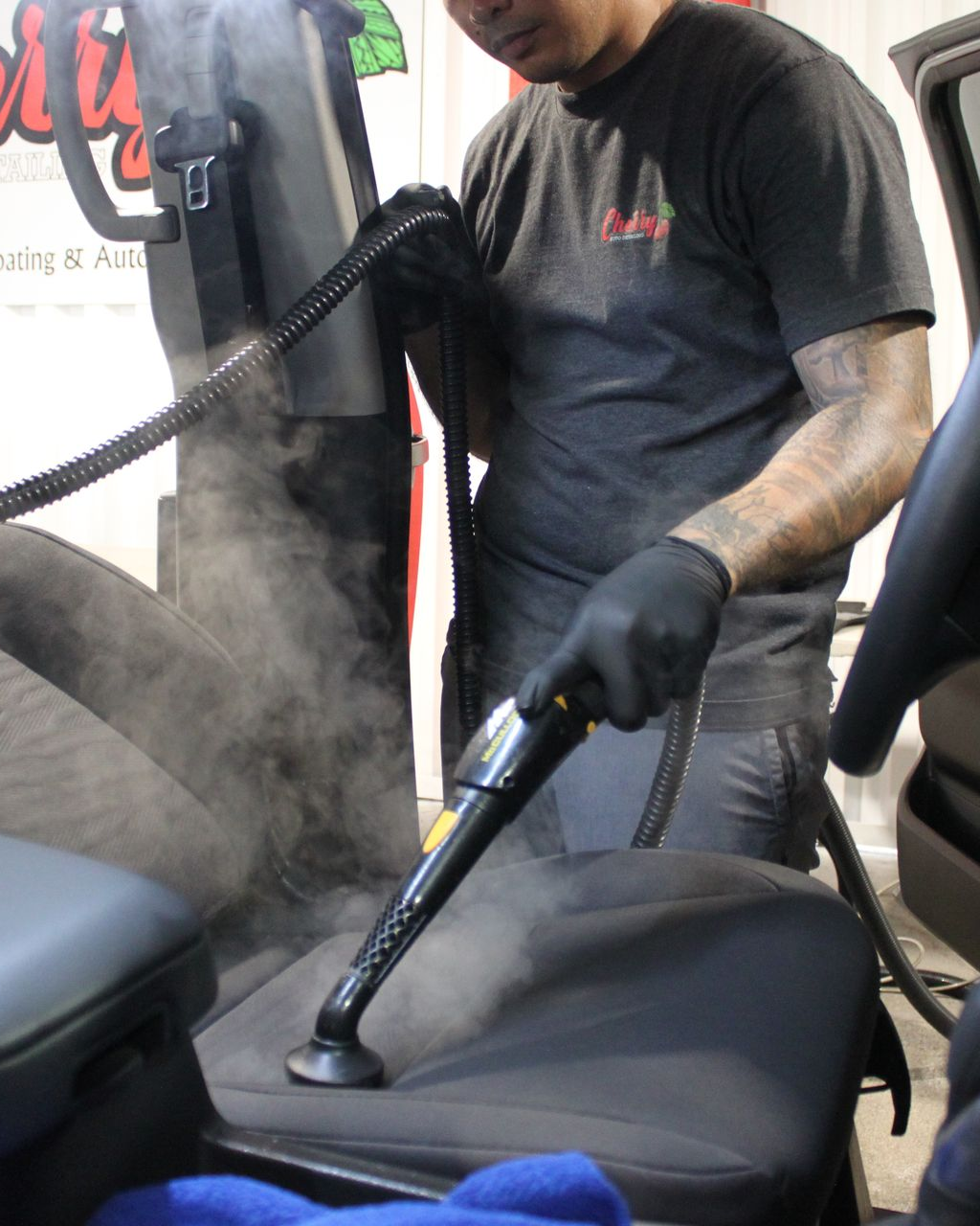 A person wearing black gloves uses a steam cleaning tool on a gray car seat inside a vehicle.