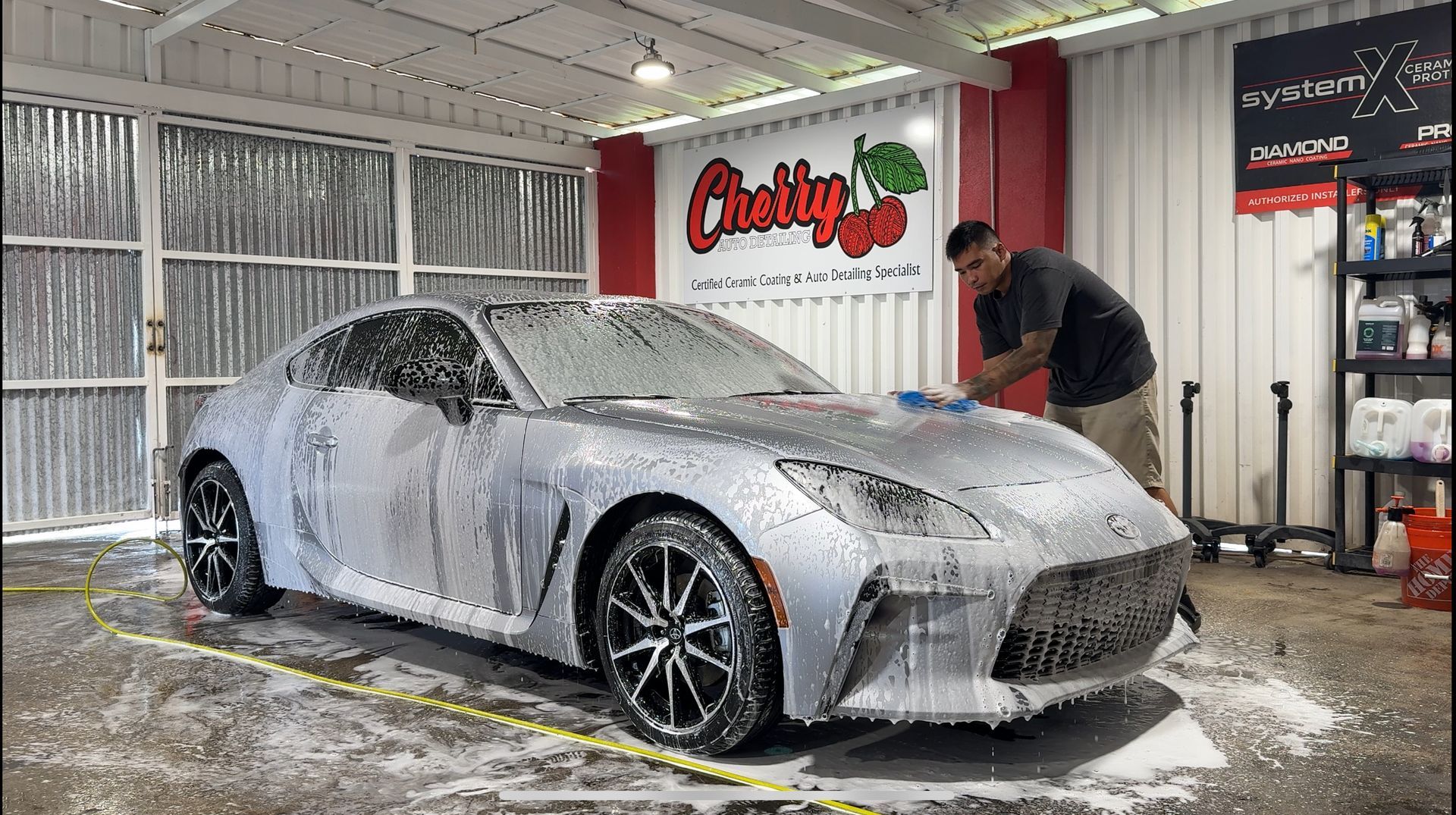 A worker hand-washes a silver car covered in white cleaning foam inside a detailing shop with a "Cherry" sign.