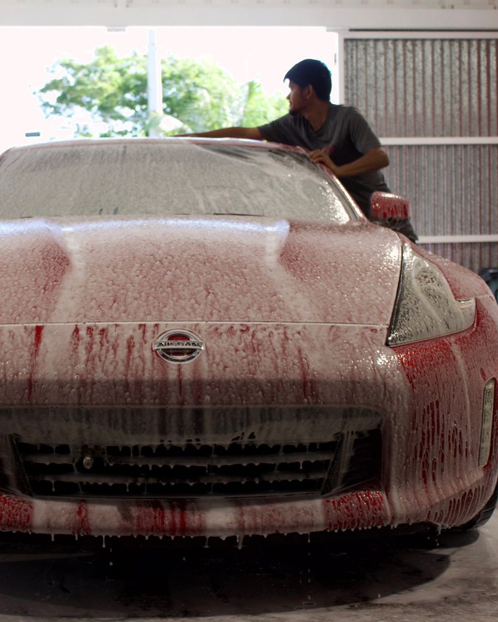 A person washing a Nissan sports car covered in red soap suds inside a garage.