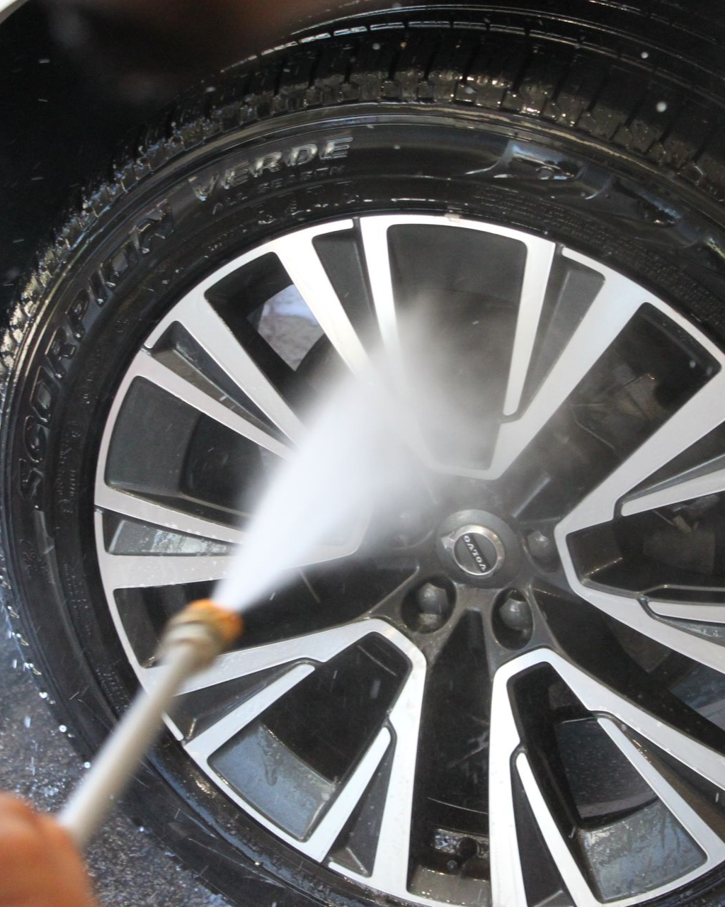 A high-pressure water sprayer cleaning a multi-spoke alloy car wheel and tire.