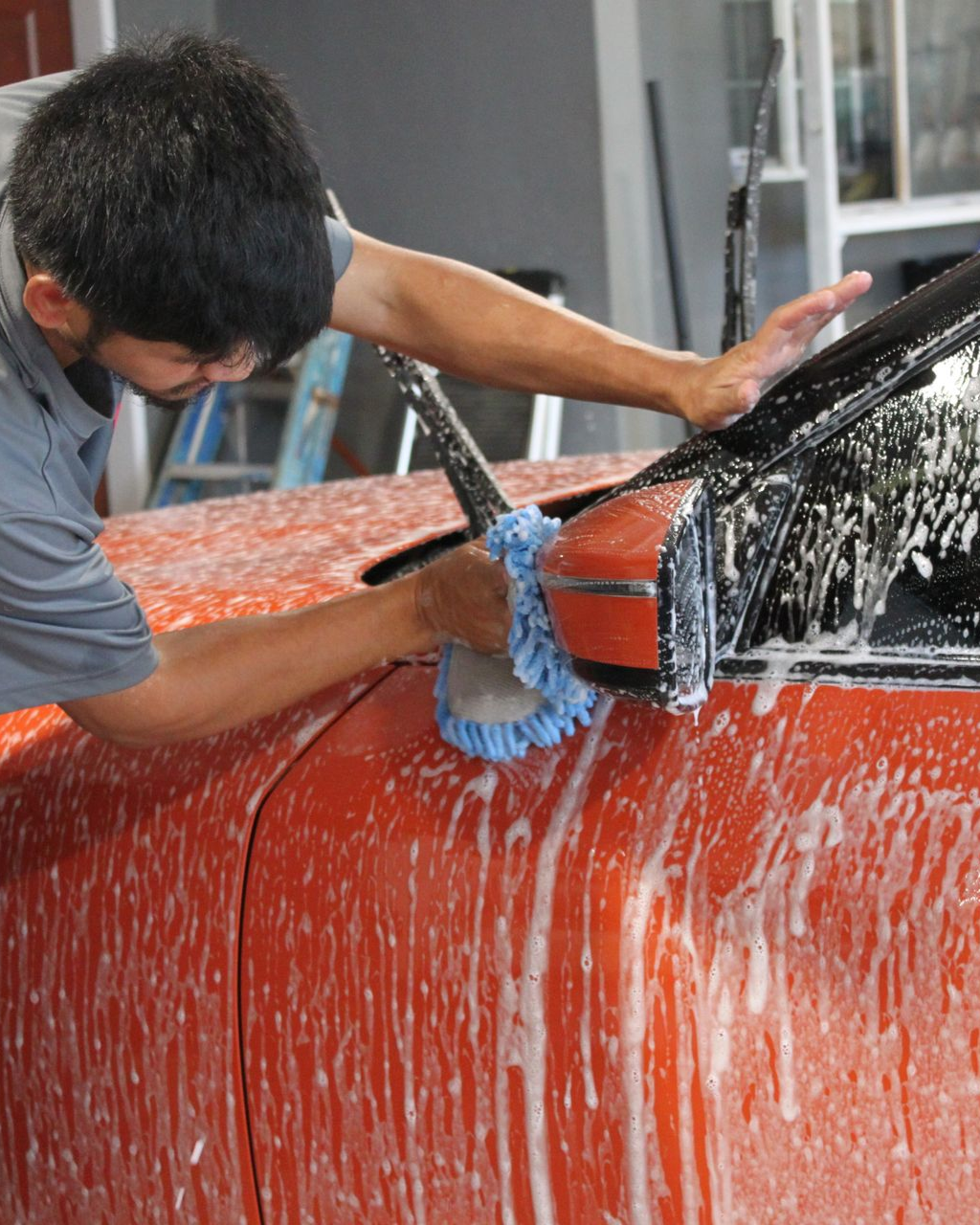 A person uses a blue microfiber mitt to wash a soapy, bright orange car, focusing on the windshield and side mirror.