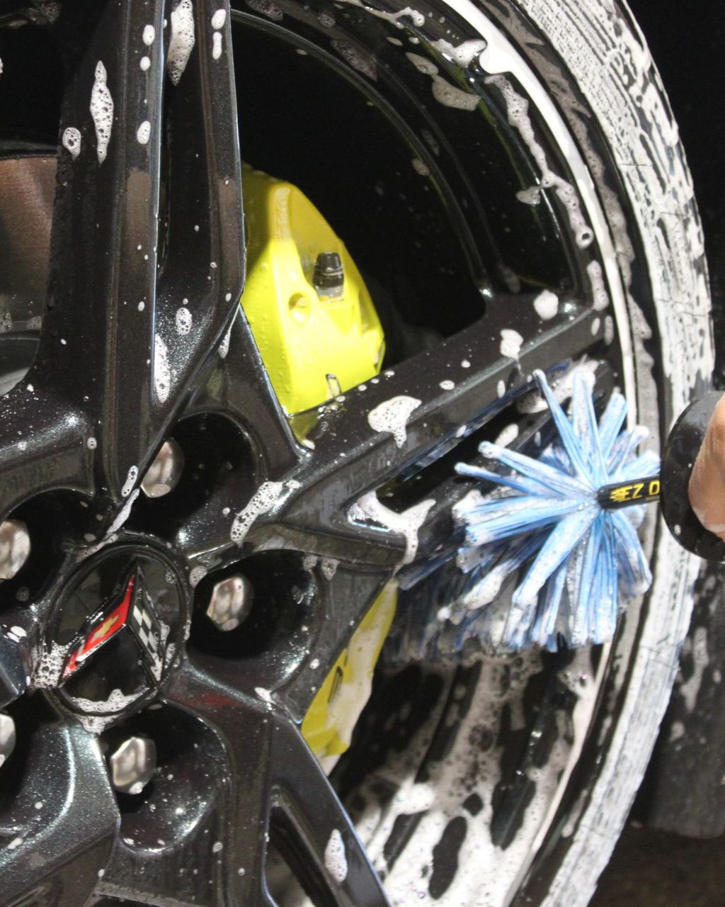 A hand uses a blue fiber brush to scrub a soapy black car wheel with a bright yellow brake caliper.
