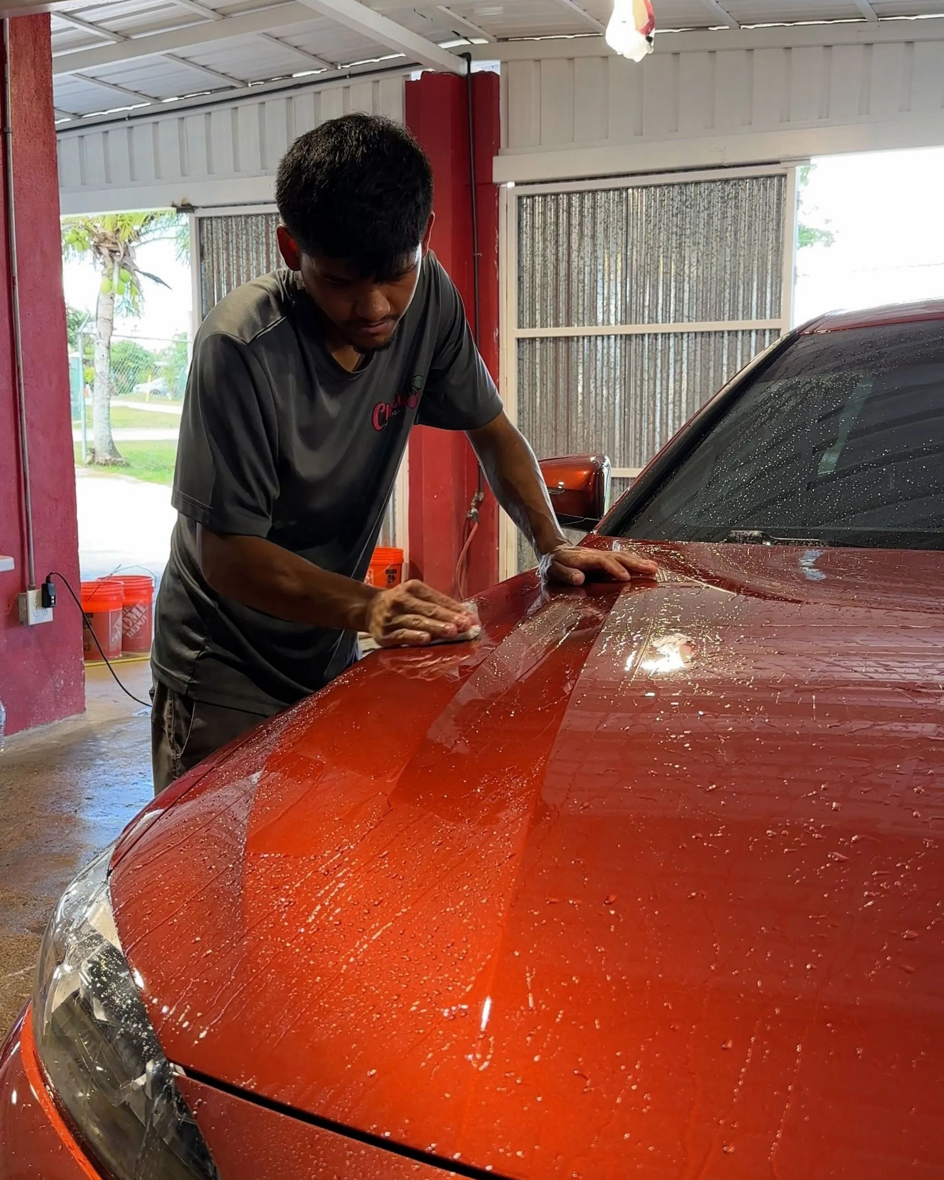 A person in a gray shirt meticulously hand-washes the wet, glossy red hood of a car inside a service bay.