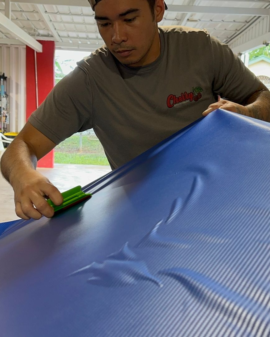 A person uses a green squeegee to smooth a blue vinyl wrap onto a surface in a workshop.