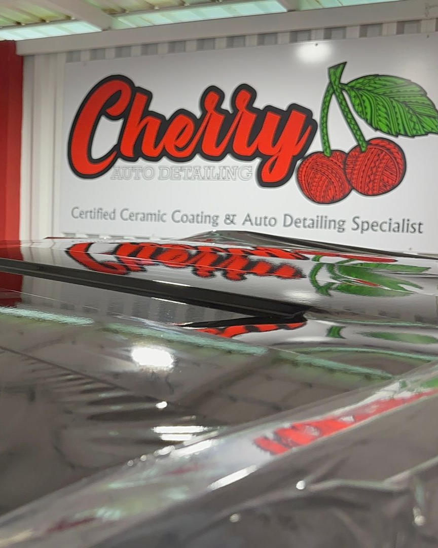 A Cherry Auto Detailing sign and logo featuring two red cherries, reflected on the glossy, dark surface of a vehicle.