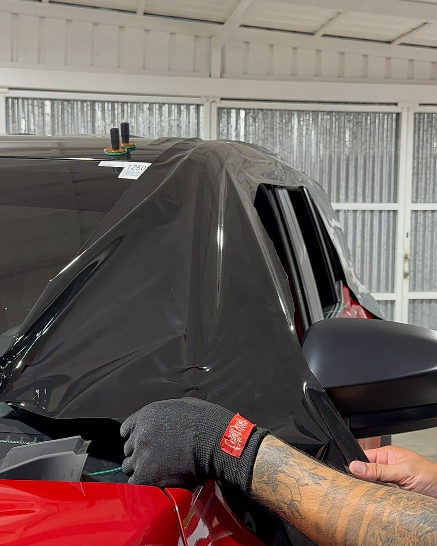 A person applying black vinyl wrap to the A-pillar and side mirror area of a red vehicle in a garage.