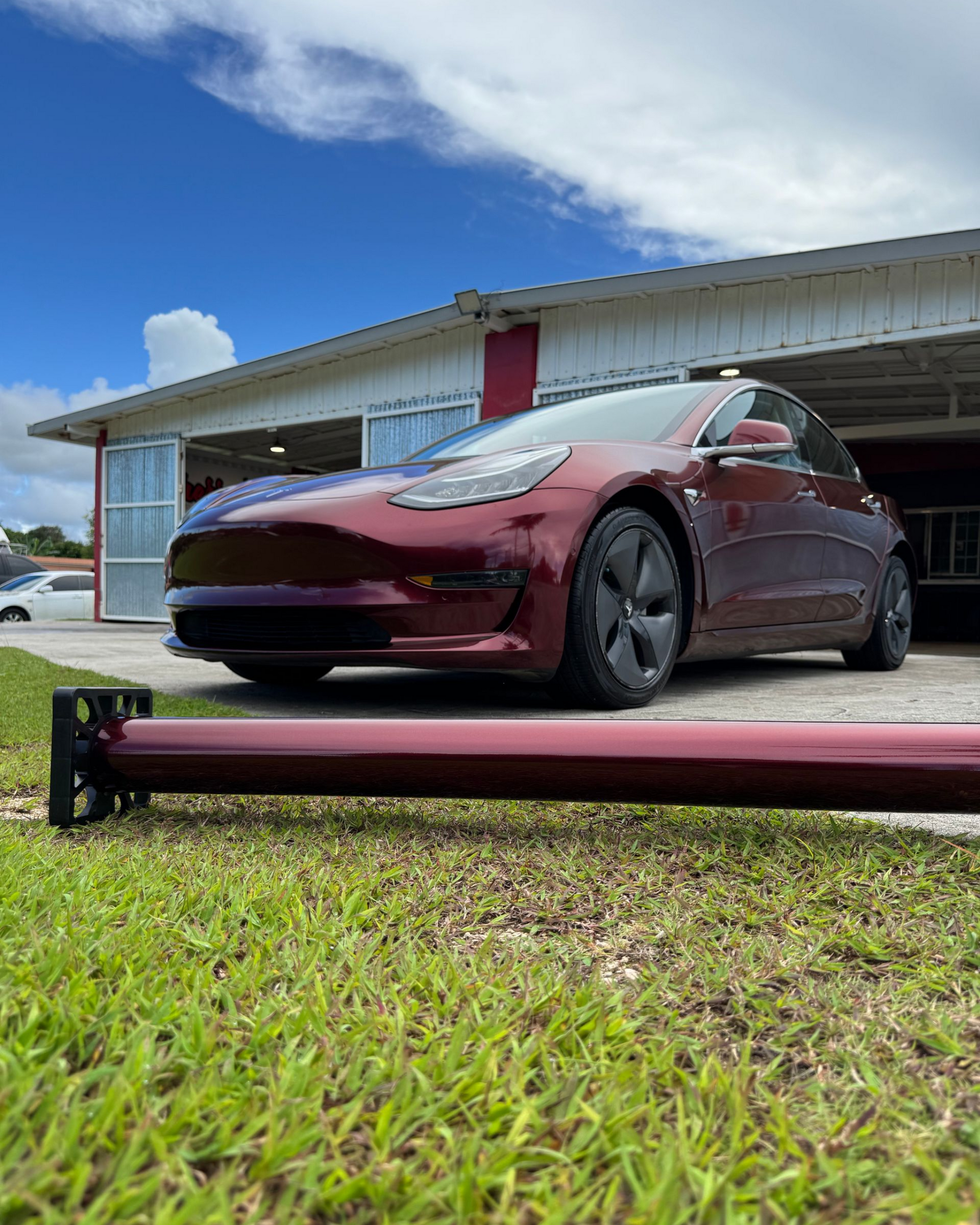 A cherry-red Tesla Model 3 sits in front of an open garage, with a matching red automotive part resting on the grass.
