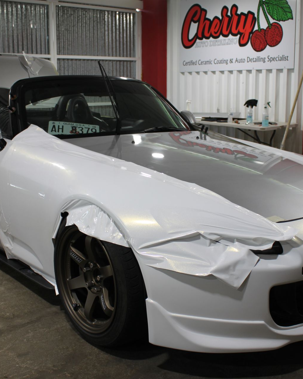 A car in a shop undergoing a white vinyl wrap, with the front fender partially covered and a cherry-themed sign behind it.