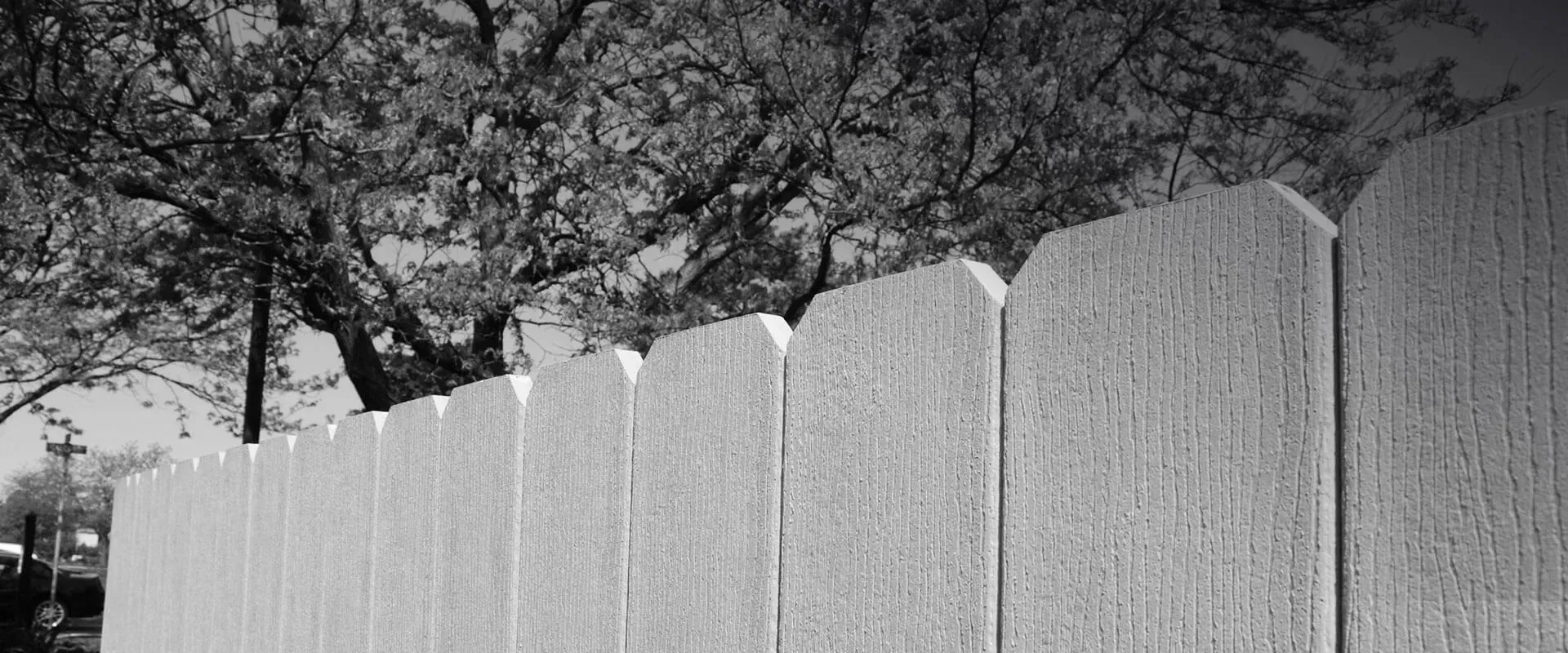 Black and white shot of a concrete fence with trees and sky in the background.