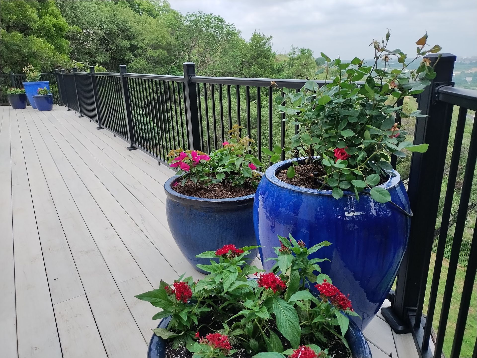 Composite deck with blue ceramic planters of flowering plants, against a railing with a scenic wooded view.
