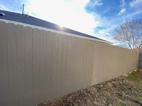 Long tan wooden fence against a blue sky, beside a home with a dark roof.