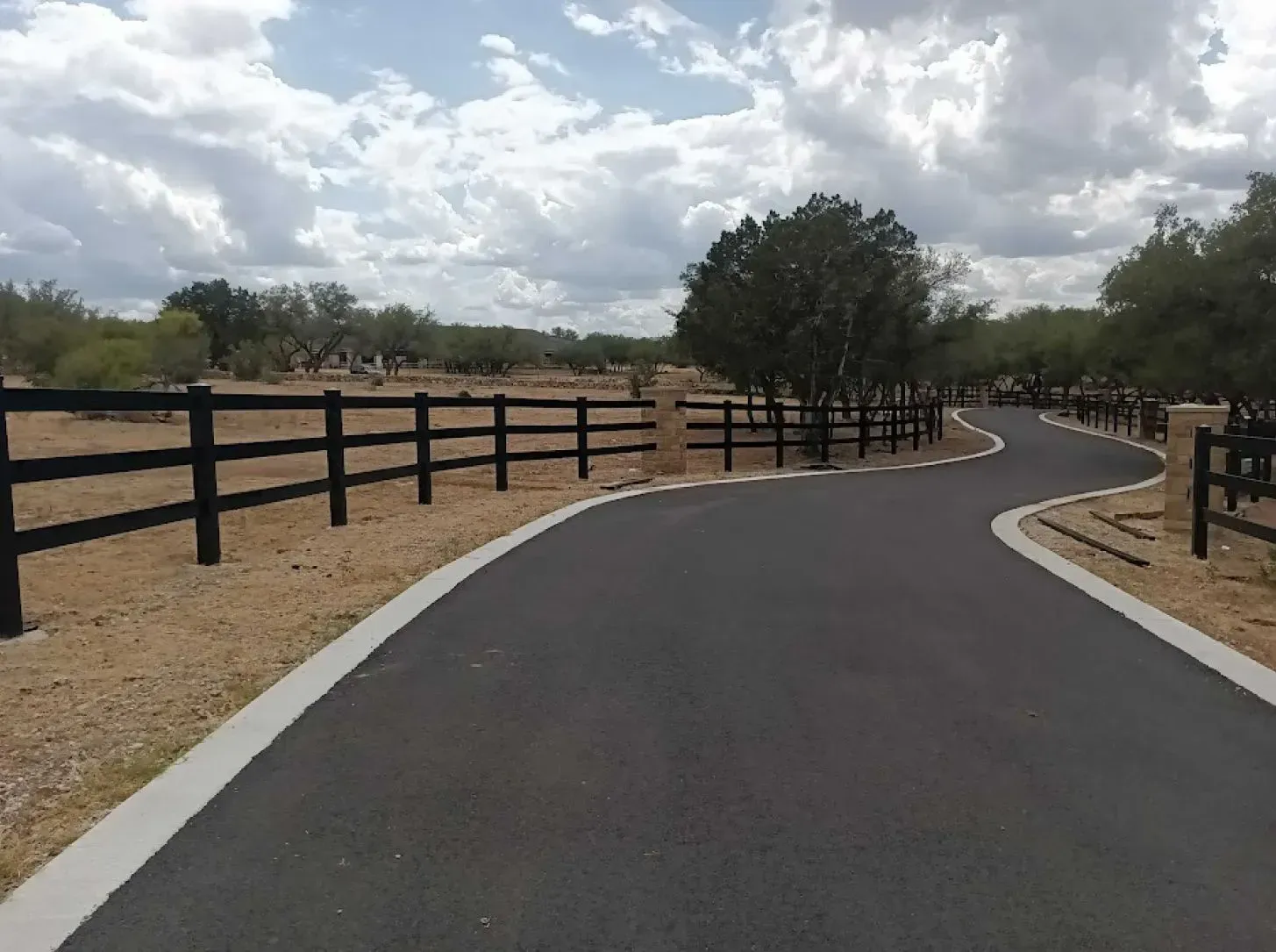 Black asphalt road curves through a rural landscape with black wooden fence and cloudy sky.