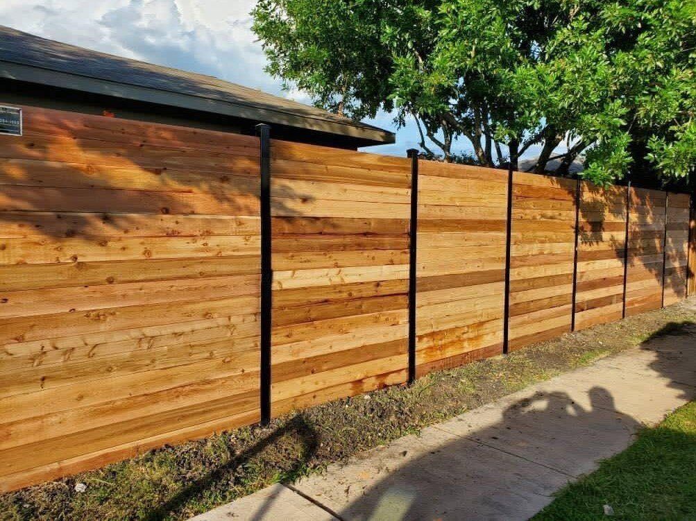 Wooden fence with black posts bordering a lawn and sidewalk, with a house in the background.