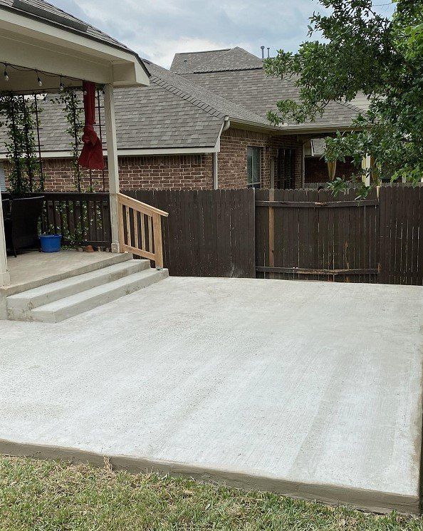 Concrete patio next to a porch with steps, wooden fence, and grassy yard.