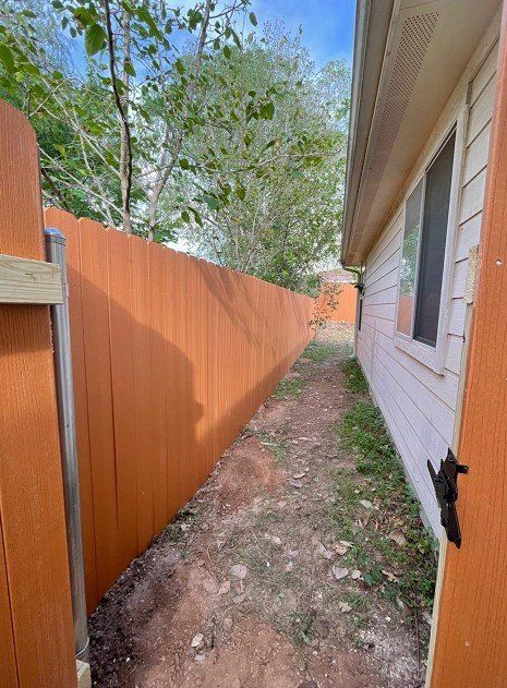 Narrow dirt path between a brown wooden fence and a white house, under a blue sky.