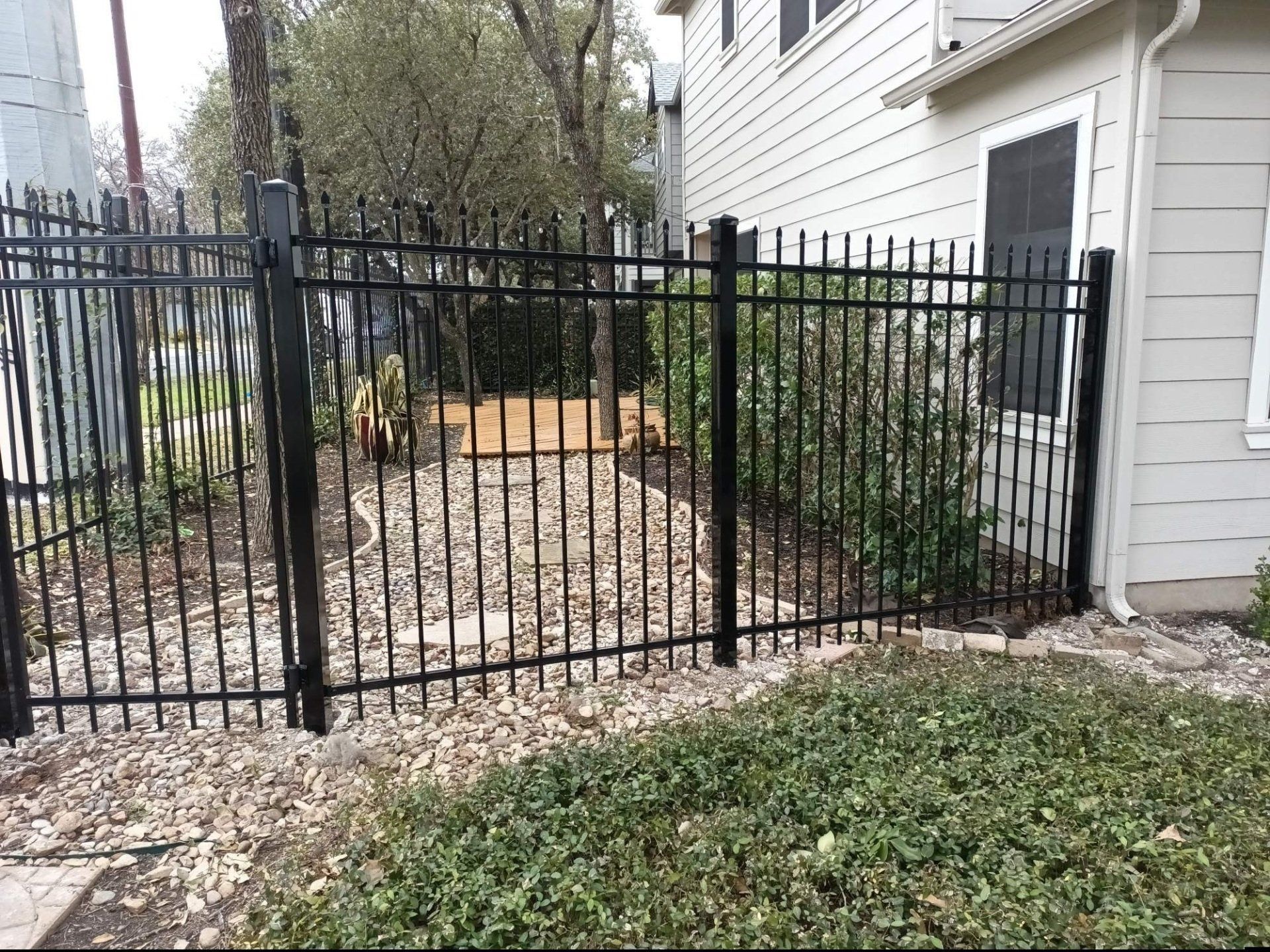 Black metal fence around a small yard with bushes and gravel, next to a house with a white exterior.