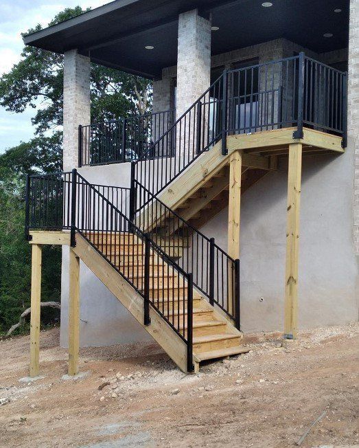 Wooden outdoor staircase with black railings, leading to a covered porch, beside a light-colored building.