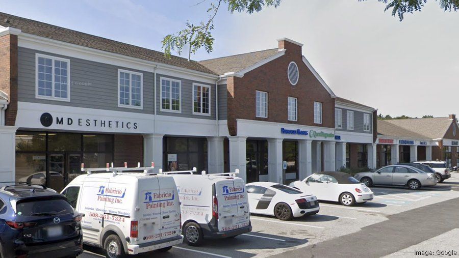 Strip mall storefronts with parked cars and vans in front under a bright sky