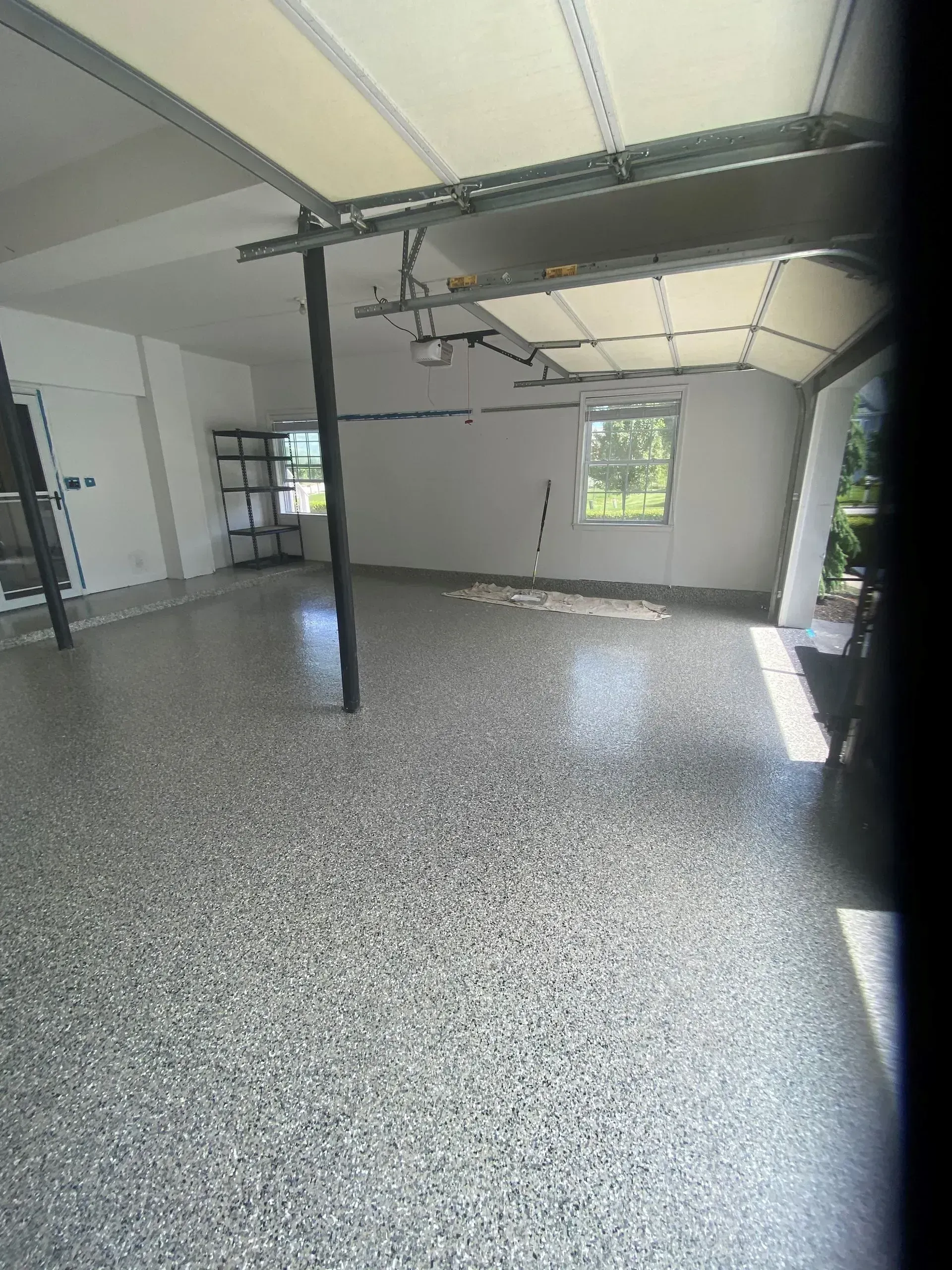 Empty garage with gray speckled floor, white walls, exposed ceiling beams, and a window at the far end