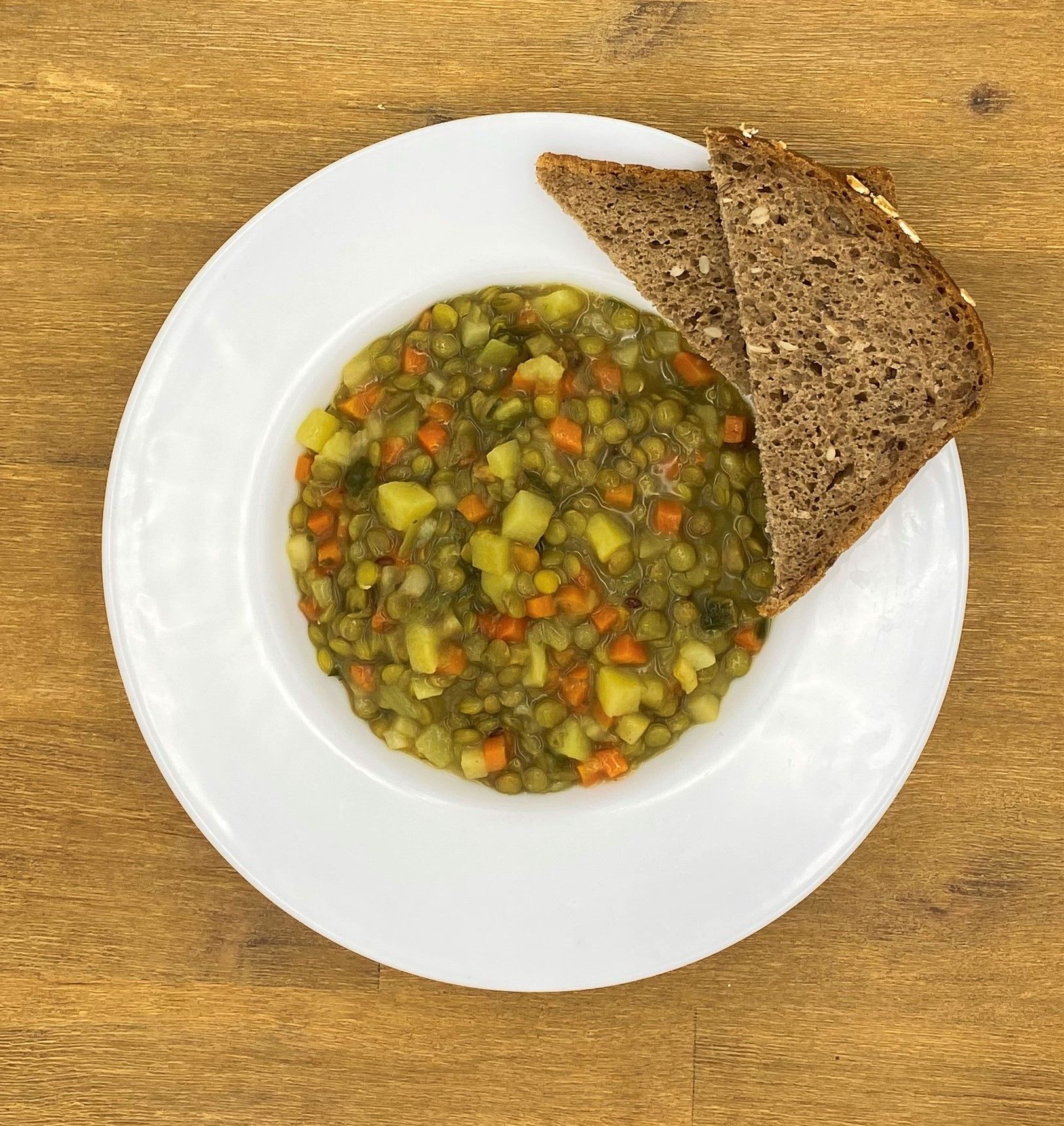 Weiße Schüssel mit Linsensuppe und Brot auf einer Holzoberfläche.