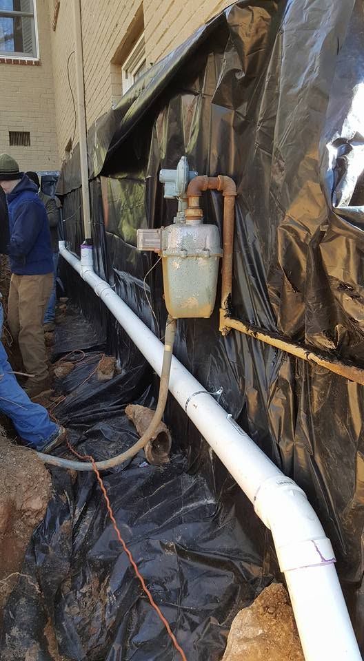 Worker beside white pipe and bucket under black tarp outside a building