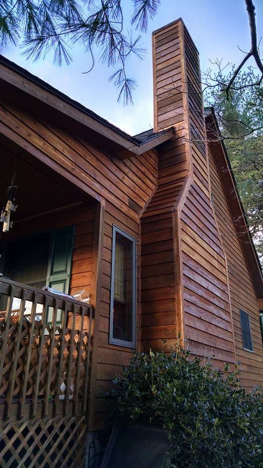 Wooden cabin exterior with a tall brick chimney and porch railing, surrounded by trees.